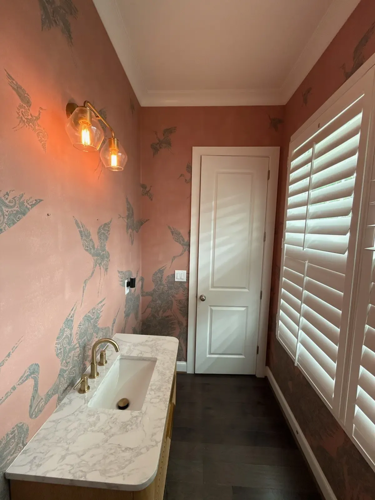 Blush wallpaper with cranes above a marble sink and brass sconce